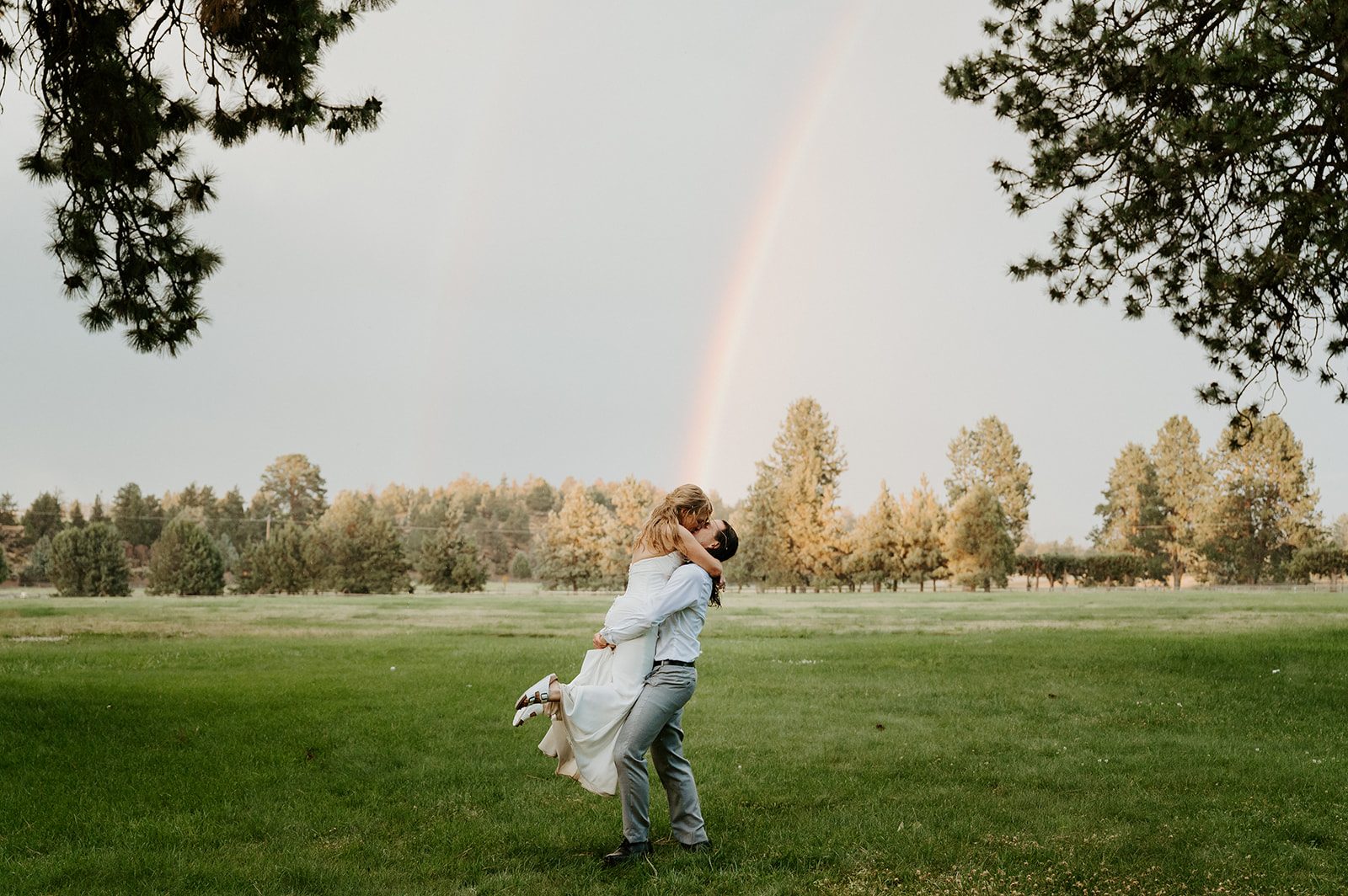 Newlyweds lifted in a kiss beneath a double rainbow in an Oregon field.