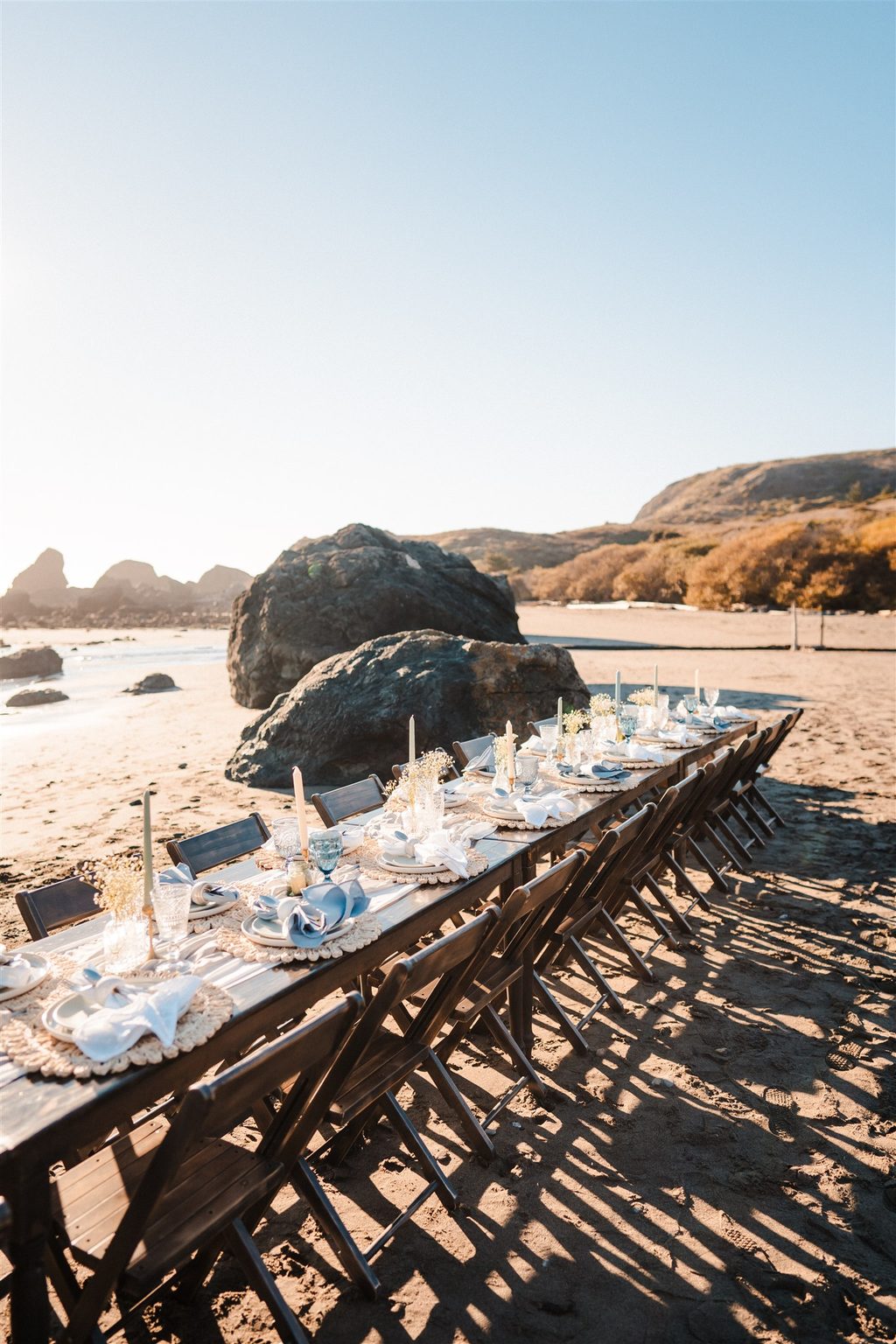 Long picnic table set with linens and florals on a sunny beach.