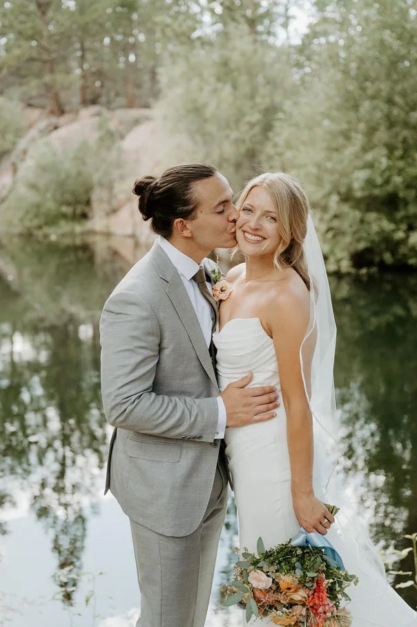 Groom kissing his bride's cheek beside a still lake, her bouquet tied with blue ribbon.